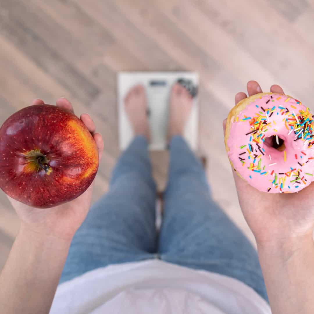 A woman standing on scales with an apple in her left hand and a iced donut in her right hand - all calories are not created equal